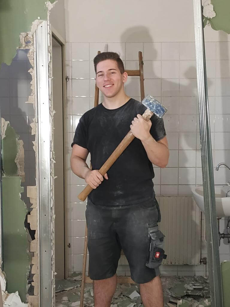 Ein junger Mann, Niklas, steht mit einem Hammer vor einer Baustelle und lächelt in die Kamera