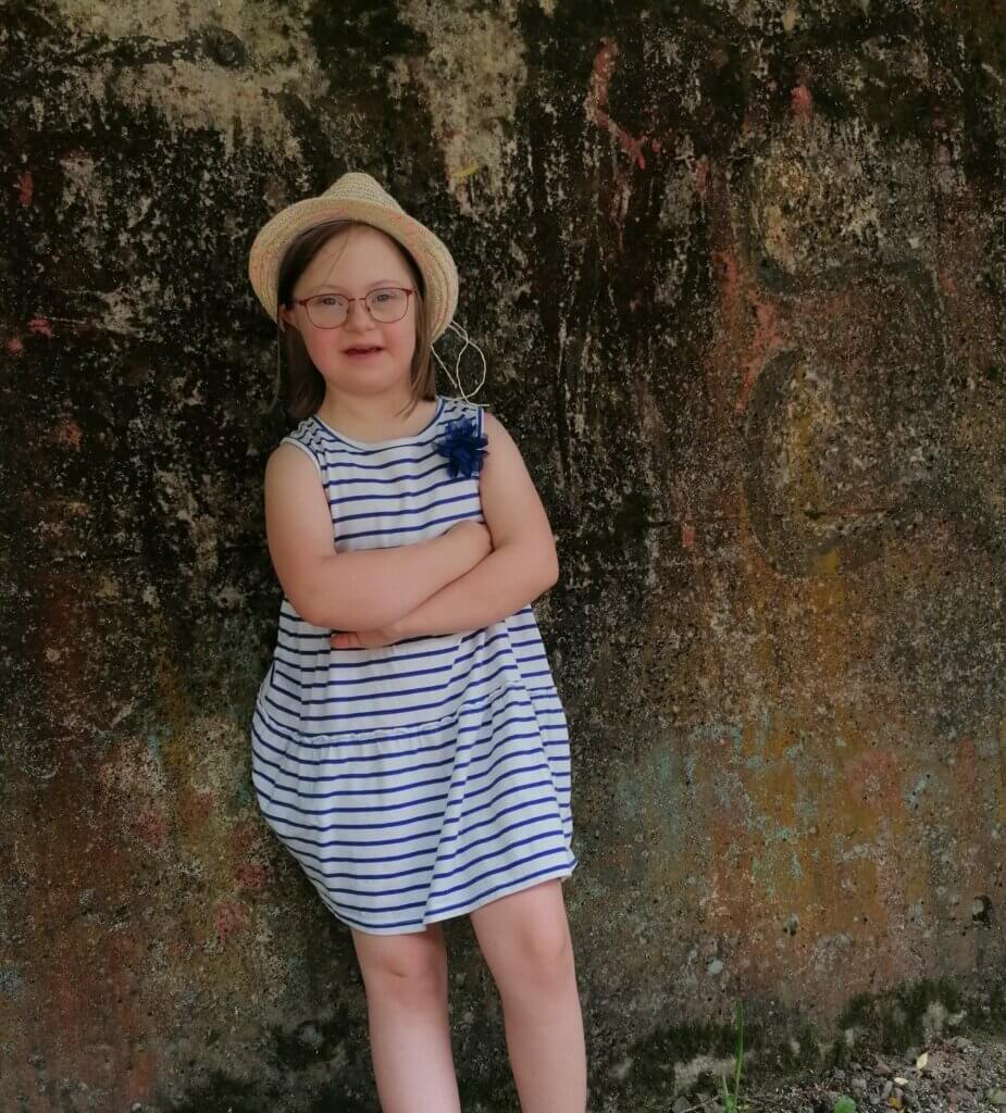 Lisa hat Trisomie 21. Sie trägt einen hellen Strohhut, ist an einen großen Baum gelehnt und hat die Arme verschränkt. sIe trägt ein blau-weiß-gestreiftes Sommerkleid und eine Brille und lächelt in die Kamera.
