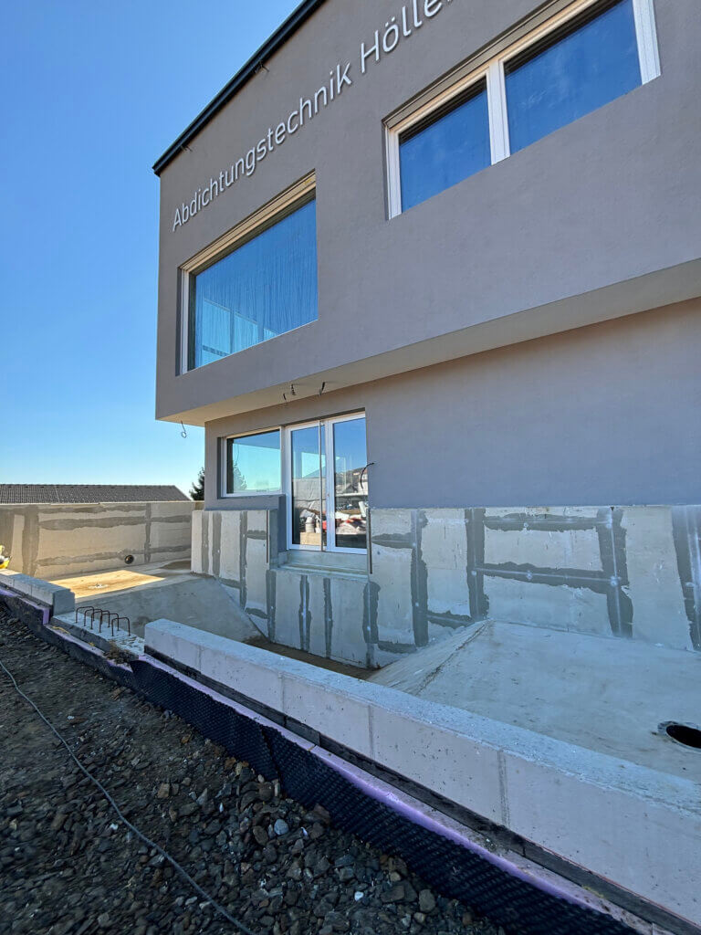 Ein Teil des Firmengebäudes ist zu sehen, die Ecke besteht aus Glas. Der obere Teil weist eine große Fensterfront auf. Davor befindet sich ein tiefer gelegenes Betonbecken. Es wird demnächst abgedichtet und sol mit Koi-Fischen gefüllt werden