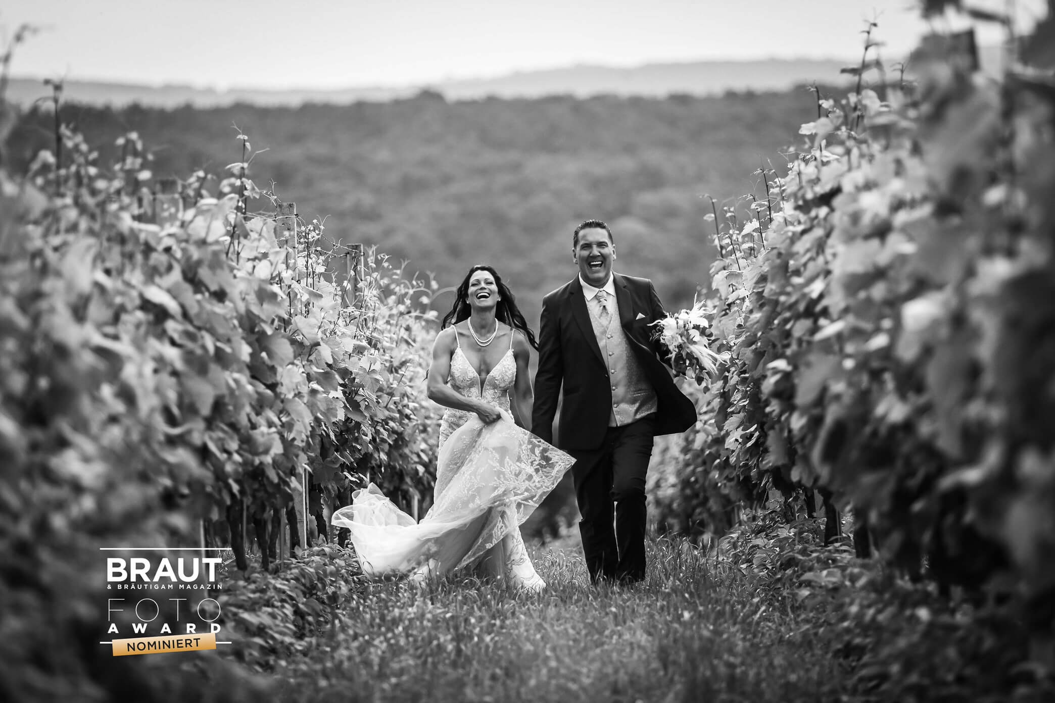 Ein Hochzeitspaar lächelt auf dem Schwer-Weiß-Bild in die Kamera. Sie sind umgeben von Weinreben.