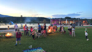 Am Greini Badesee fand das Familiencampen statt. Erwachsene und Kinder sind auf einer Wiesse zu sehen kurz nach Sonnenuntergang mit Lagerfeuer.