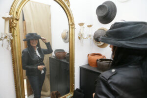 Trude Meichenitsch vor dem Spiegel. Sie blickt hinein und trägt einen schwarzen Hut