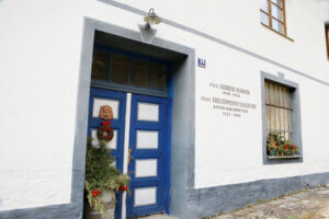 Die Eingangstür des Hauses von Trude Meichenitsch im Großformat. Die Tür ist blau-weiß. Links davon steht eine grüne Winterpflanze. Recht ist die Aufschrift "ehem. Gerberei Schiefer 1838-1956 ehem. Edelserpentin Schleiferei Anton Eichenitsch 1957-1995