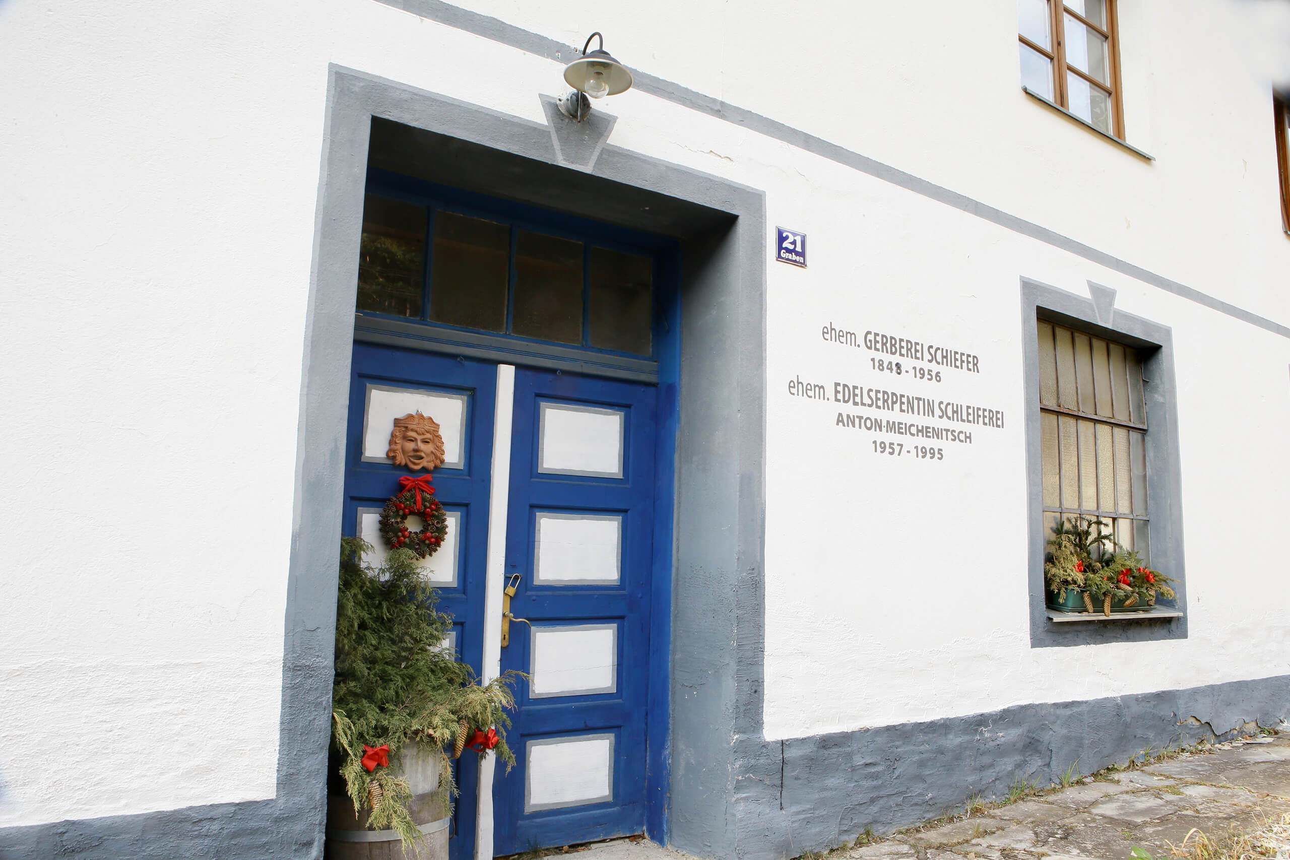 Die Eingangstür des Hauses von Trude Meichenitsch im Großformat. Die Tür ist blau-weiß. Links davon steht eine grüne Winterpflanze. Recht ist die Aufschrift "ehem. Gerberei Schiefer 1838-1956 ehem. Edelserpentin Schleiferei Anton Eichenitsch 1957-1995
