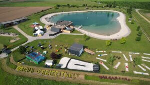 Die Greini Freizeit-Welt ist von oben zu erkennen. Der Badesee mit den vielseitigen Anlagen (Beachvolleyball, Minigolf, Trampolin) ist zu erkennen. Der BAdesee ist von einer grünen Wiese umgeben.