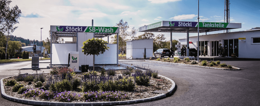 Tankstelle Stöckl von ausßen. Logo violet und grün. Von außen fotografiert mit Blumenbeet als Umfahrung und Waschanlage etc.