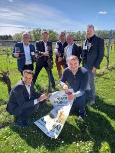 Sieben Männer halten Weingläser in der Hand und präsentieren das Plakat des Weinfrühlings 2024.