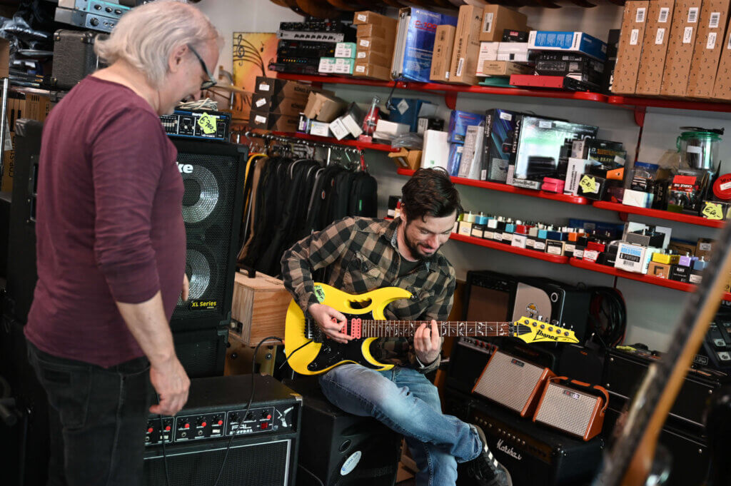 Manuel Kants hält eine gelbe E-Gitarre in der Hand und spielt. Neben ihm steht Helmut Lang und sieht ihm dabei zu. Im Hintergrund ist erkennbar, dass sich beide in der Werkstatt befinden.