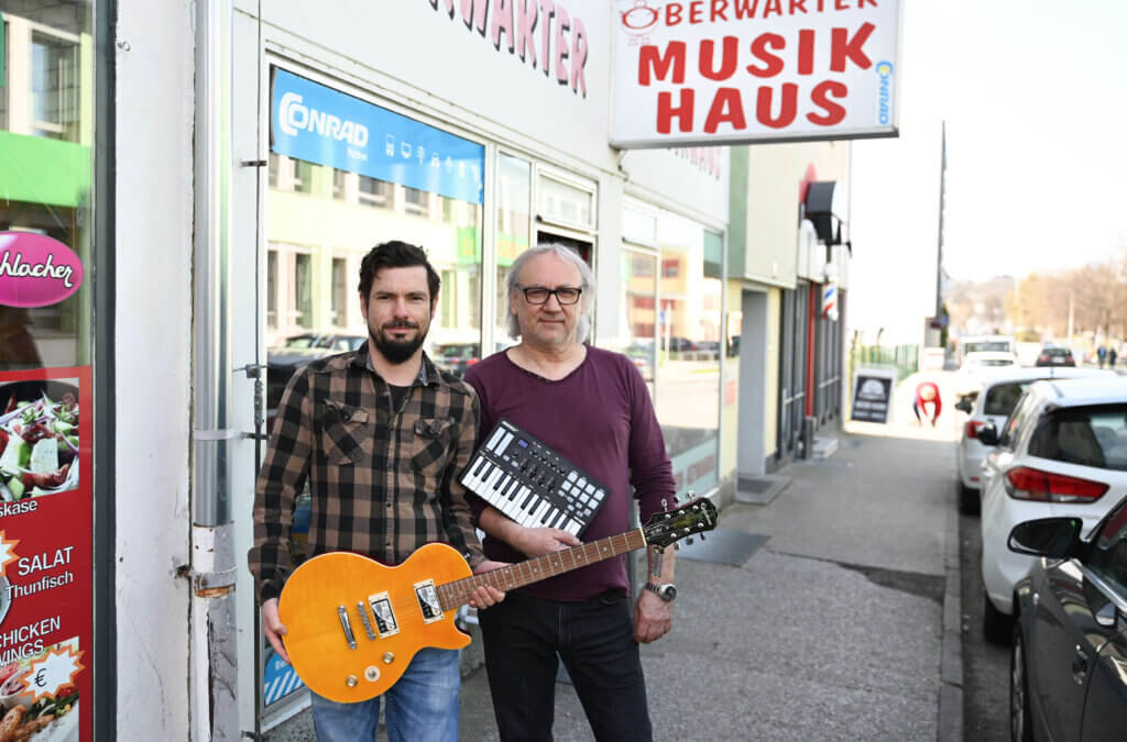 Manuel Kants und Helmut Lang stehen vor dem Musikhaus Oberwart. Rechts oben im Bild sieht man die Reklametafel. Manuel hält eine Gitarre in der Hand. Helmut ein Mini-Keyboard
