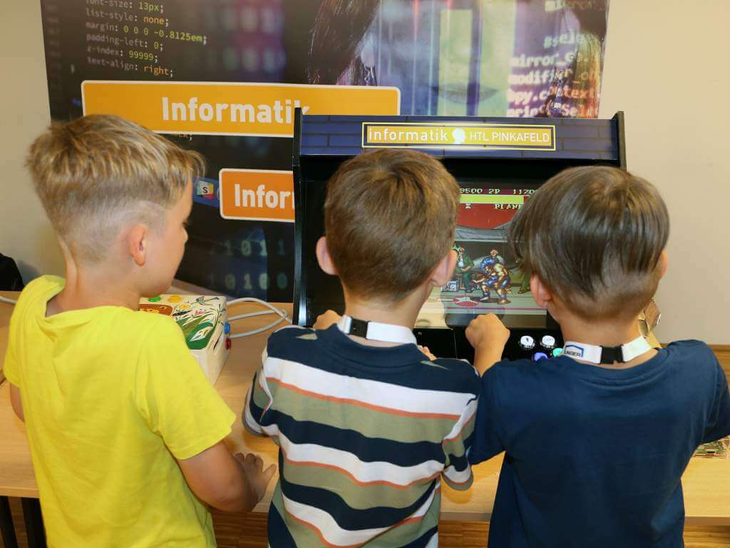 Drei Kinder blicken in einen Computer. Li ks davon sieht man die Aufschrift Informatik