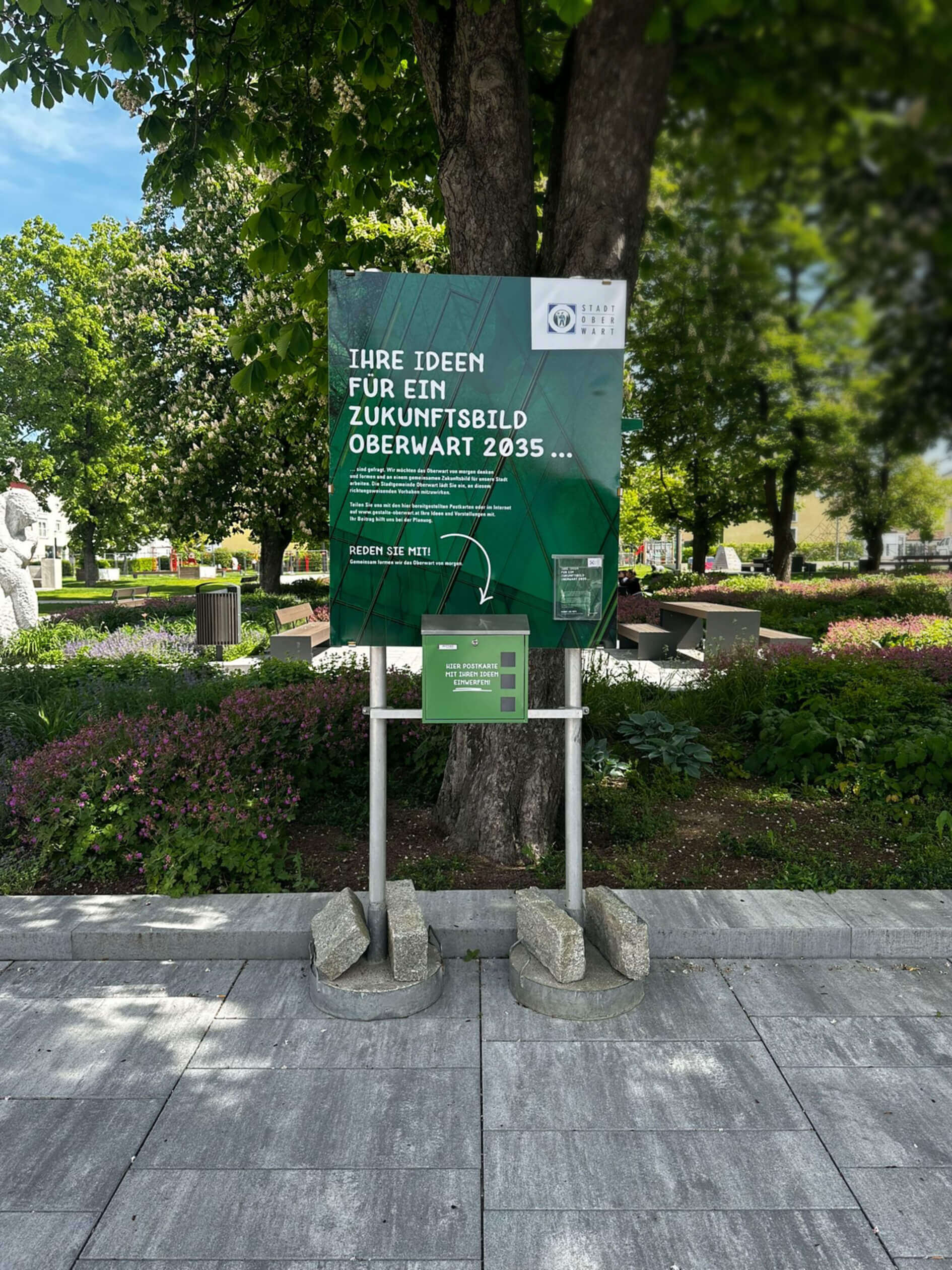 Auf diesem Bild sieht man eine Tafel mit einem Briefkasten die sich im Stadtgarten in Oberwart befindet. Die tafel ist grün mit weißer Schrift und darauf steht "Ihre Ideen für ein Zukunftsbild Oberwart 2035..."