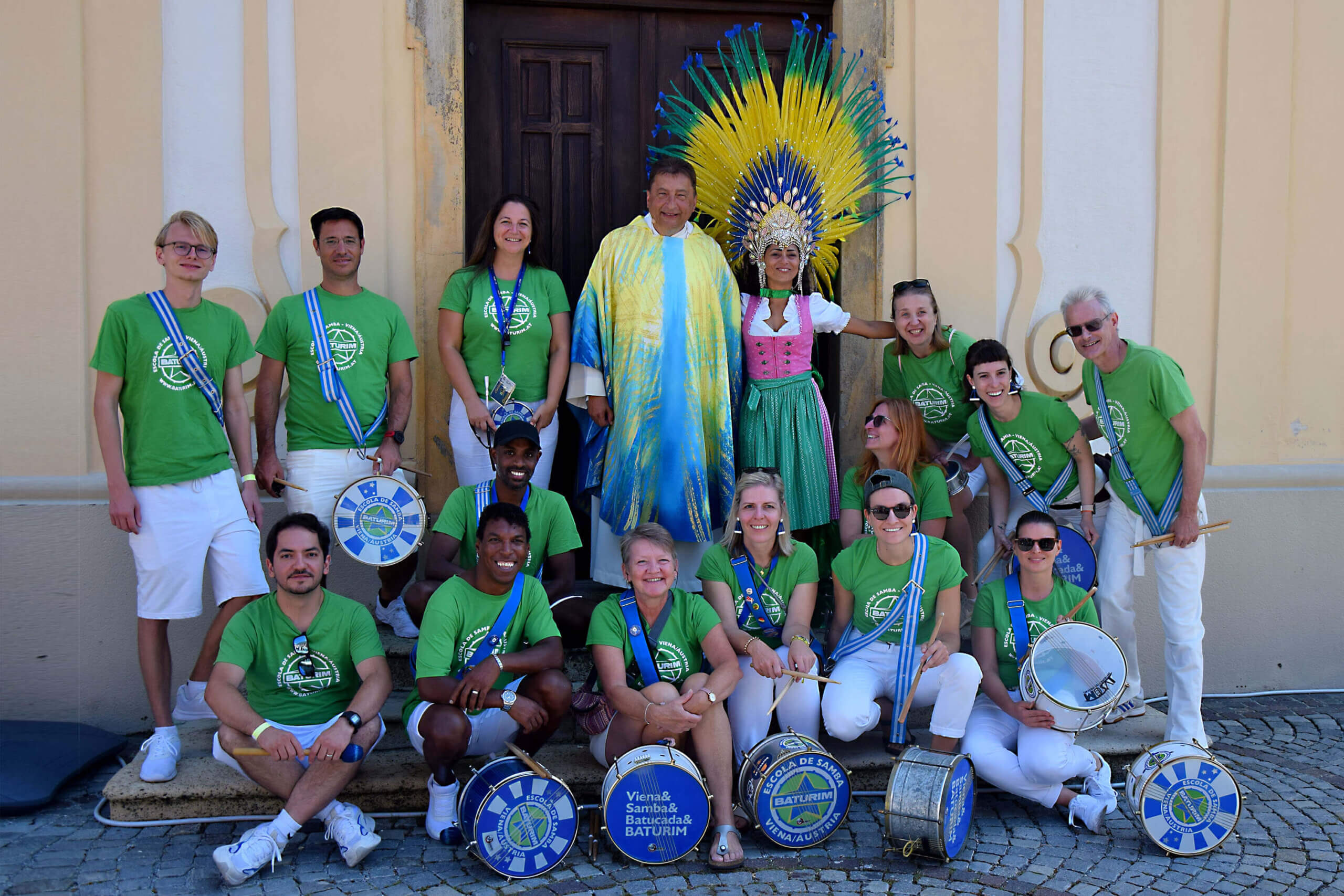 Eine Gruppe von etwa fünfzehn Personen posiert fröhlich vor einem Gebäude. Die meisten tragen grüne T-Shirts mit weißen Hosen oder Shorts. Ein Mann in der Mitte trägt ein farbenfrohes Gewand, und eine Frau daneben trägt ein Dirndl mit einem auffälligen, gelben und blauen Kopfschmuck. Mehrere Mitglieder der Gruppe halten Trommeln oder andere Musikinstrumente.