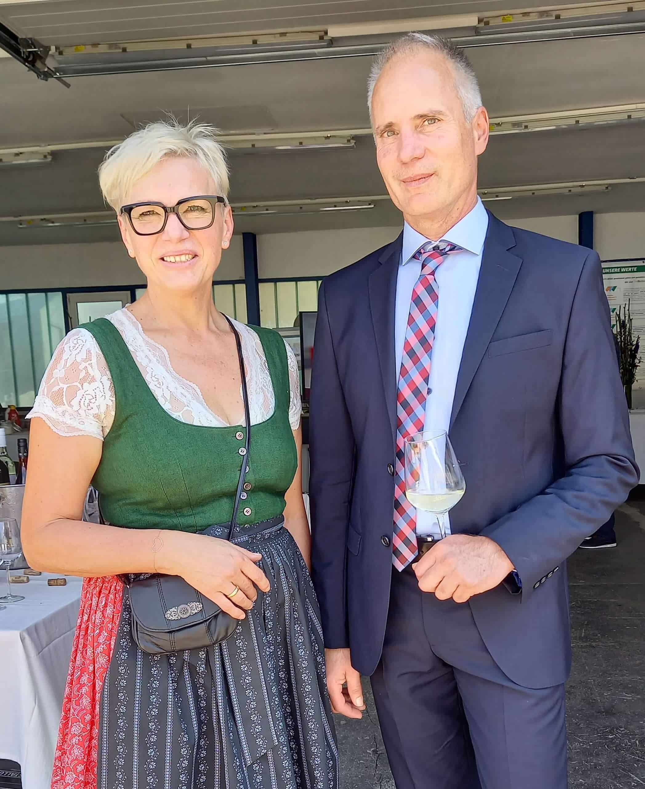Das Bild zeigt Dr. Mag. David Reiter und seine Gattin Johanna, die in einem offenen, industriellen Umfeld stehen, während der 25-Jahres-Feier. Dr. Reiter ist ein Mann mittleren Alters und trägt einen dunkelblauen Anzug mit einem hellblauen Hemd und einer rot-blau gestreiften Krawatte. Er hält ein Weinglas in der rechten Hand. Seine Haltung ist aufrecht und sein Gesichtsausdruck freundlich. Johanna steht neben ihm und trägt ein traditionelles Dirndl, das aus einer grünen Bluse mit Spitzenbesatz und einem zweifarbig gemusterten Rock besteht, dessen Hauptfarben Dunkelblau und Rosa sind. Sie trägt eine kleine, dunkle Umhängetasche und schwarze flache Schuhe. Ihre Haare sind kurz und blond, und sie trägt eine Brille mit runden Gläsern. Beide schauen direkt in die Kamera und lächeln. Der Hintergrund zeigt Teile eines Zeltes oder einer ähnlichen Struktur, die auf ein Event im Freien hindeutet.