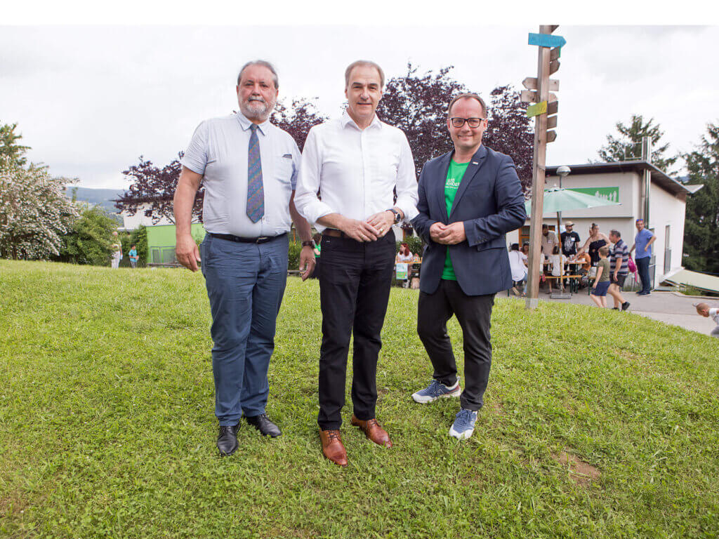 Auf dem Bild stehen drei Männer entspannt auf einer Wiese vor dem Hintergrund des SOS Kinderdorfs in Pinkafeld. In der Mitte steht Soziallandesrat  Leonhard Schneemann, rechts von ihm Kinderdorf-Leiter Marek Zeliska und links Bezirkshauptmann Peter Bubik. Alle drei tragen lässige Geschäftskleidung: Schneemann und Bubik in weißen Hemden und dunklen Hosen, während Zeliska ein grünes SOS-Kinderdorf-Shirt unter seinem dunklen Blazer trägt. Im Hintergrund ist ein Teil des Festes sichtbar, mit Menschen, die sich in einer entspannten Atmosphäre im Freien aufhalten. Die grüne Umgebung und der informelle Rahmen des Treffens spiegeln die familiäre und unterstützende Umgebung des SOS Kinderdorfs wider.
