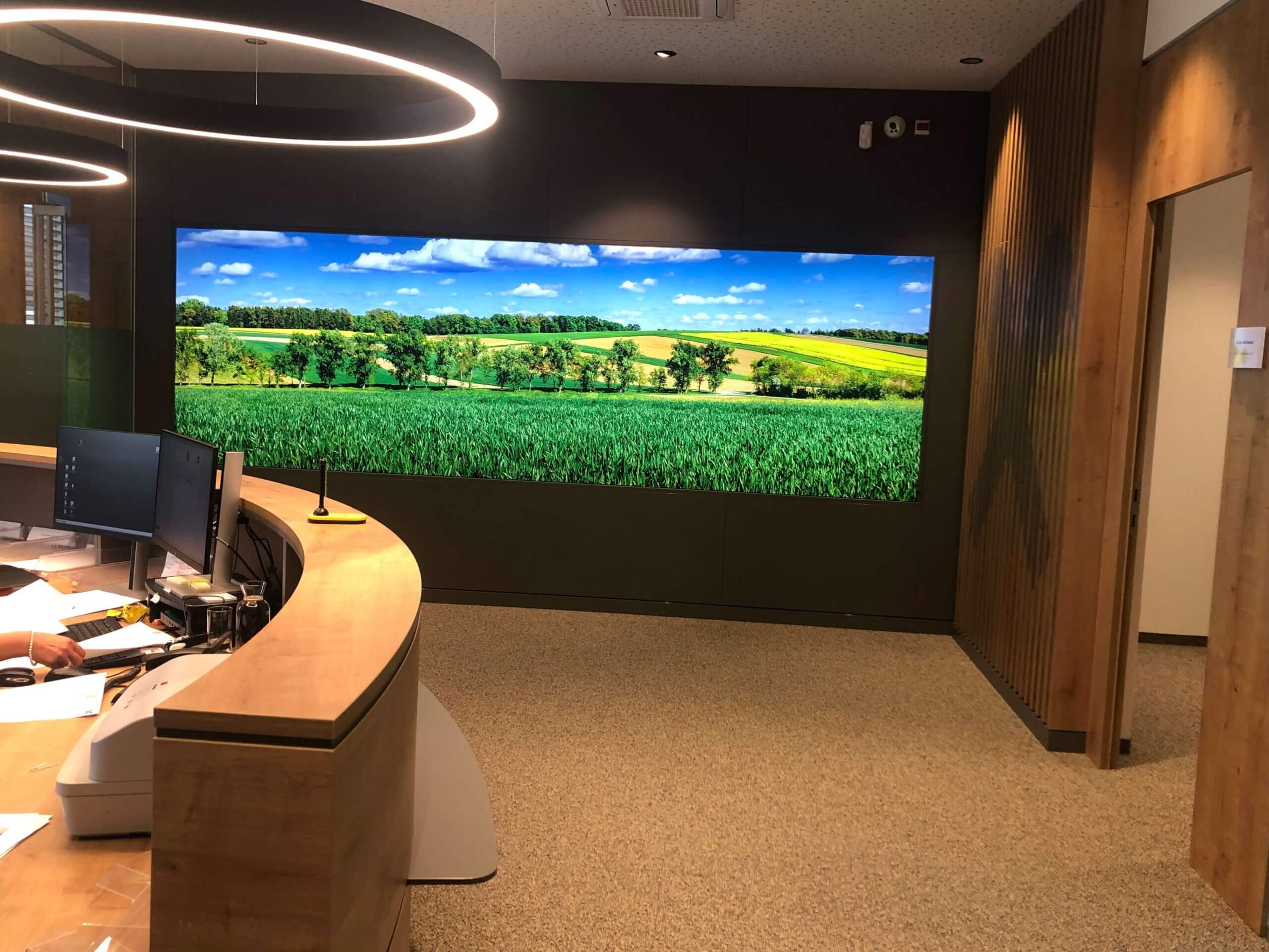 Empfangsbereich der erweiterten Raiffeisenbank Oberwart. Ein großer, gebogener Holztresen mit mehreren Computerarbeitsplätzen ist zu sehen. Im Hintergrund hängt ein großes Bild einer grünen Landschaft mit Feldern und Bäumen unter einem blauen Himmel an der Wand.