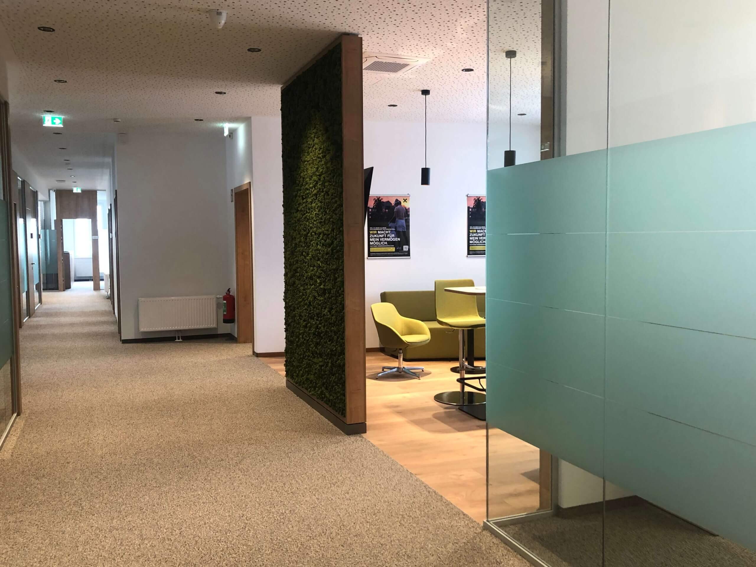 Ein Flur in der Raiffeisenbank Oberwart. Rechts ist eine Wand mit einer grünen Moosverkleidung. Links befinden sich Glastrennwände mit einem matten Streifenmuster. Im Hintergrund steht eine Sitzgruppe mit hellgrünen Sesseln und einem runden Tisch.