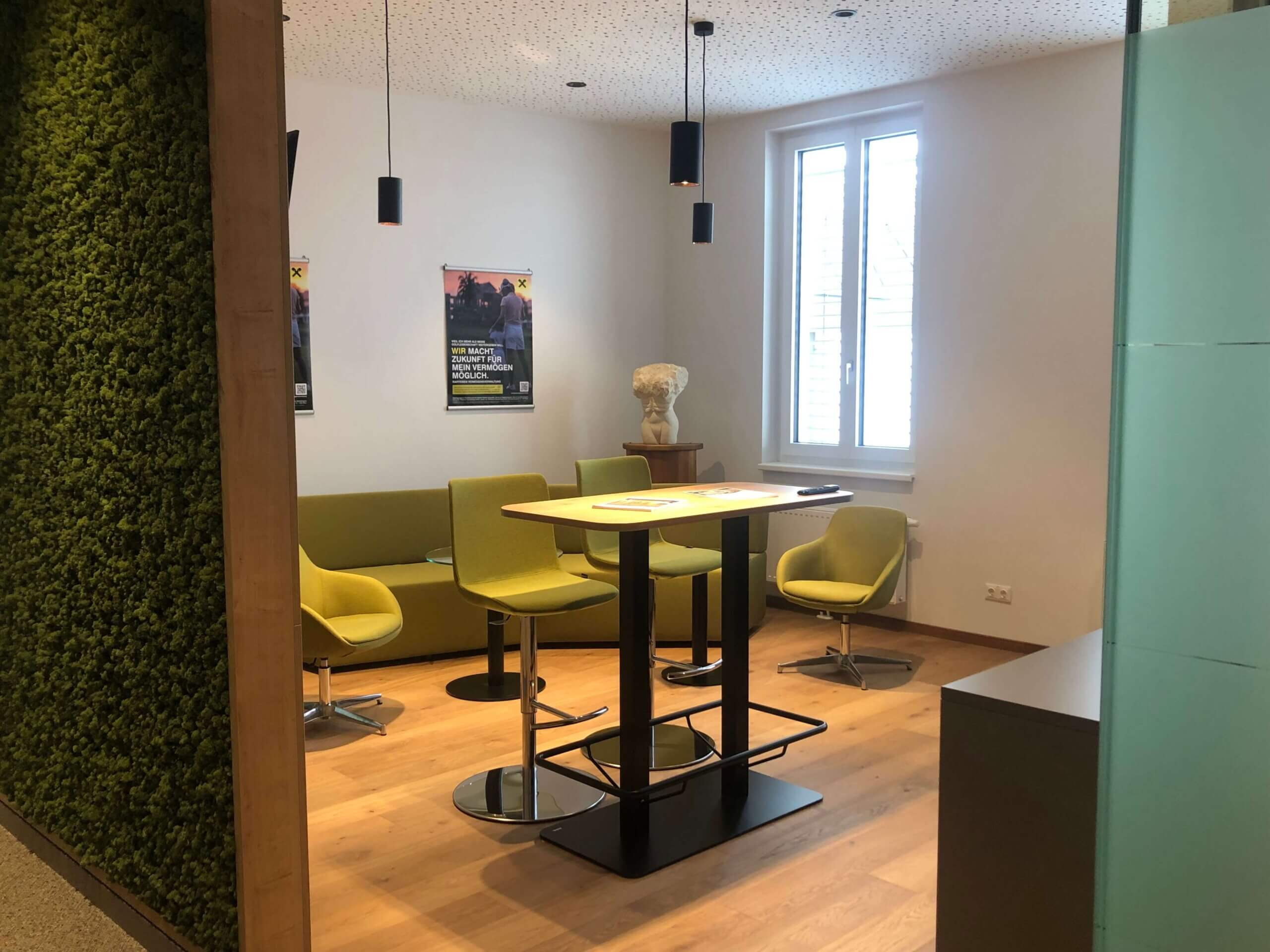 Eine gemütliche Sitzecke in der Raiffeisenbank Oberwart. Die Wände sind weiß, die Decke hat kleine, punktuelle Beleuchtungen. In der Ecke steht eine grüne Sitzbank mit dazu passenden Sesseln und einem hohen Tisch. An der Wand hängen zwei Poster.
