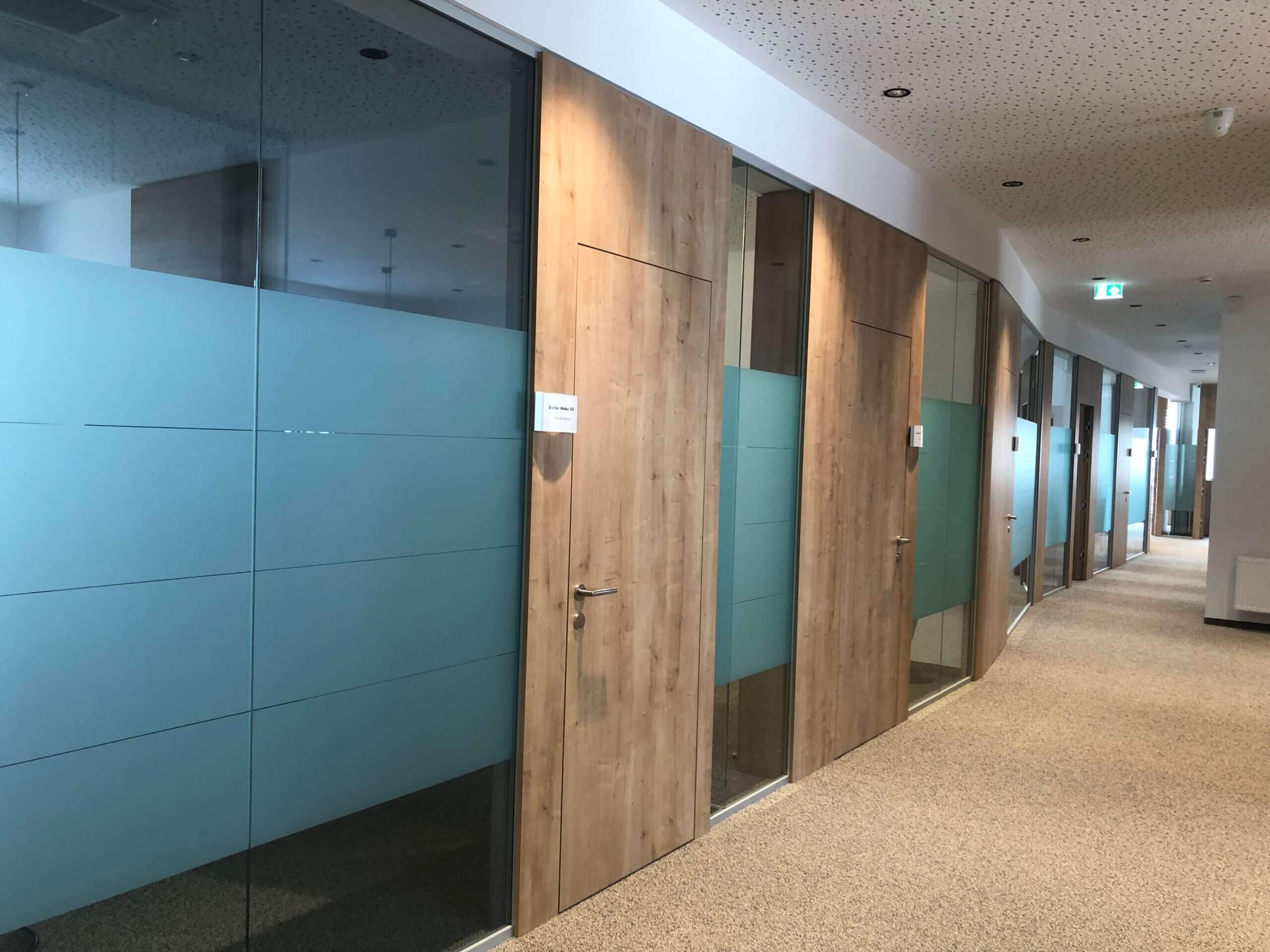 Ein langer, heller Flur mit Büros in der Raiffeisenbank Oberwart. Links sind Büros mit Glastüren und Holzelementen. Der Boden ist mit Teppich ausgelegt, und die Decke ist mit kleinen, punktuellen Leuchten versehen.