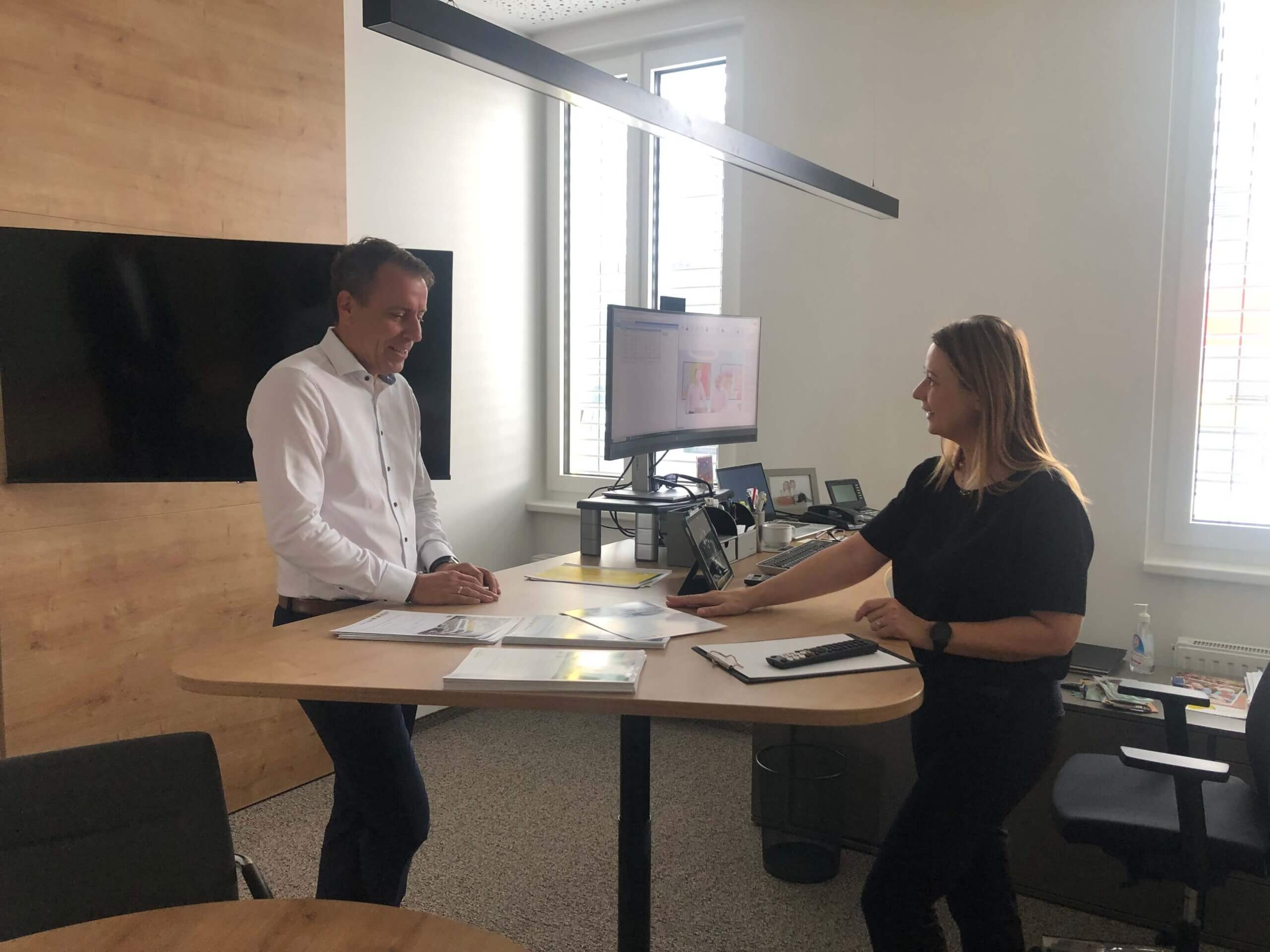 Ein moderner Büroraum mit zwei Personen in der Raiffeisenbank Oberwart. Eine Frau und ein Mann stehen an einem hohen Schreibtisch und besprechen Dokumente. Im Hintergrund ist ein Fenster mit Jalousien, und an der Wand hängt ein großer Flachbildfernseher.