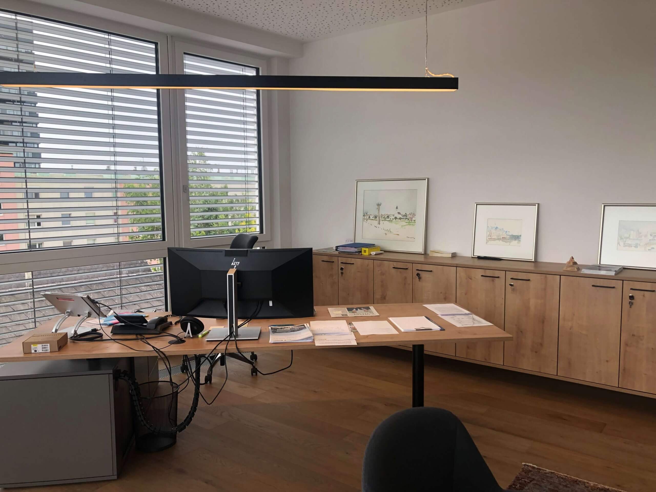 Ein weiteres Büro in der Raiffeisenbank Oberwart. Ein großer Schreibtisch mit Computer und Büromaterialien steht vor bodentiefen Fenstern mit Jalousien. Die Rückwand des Büros ist mit Holzmöbeln und mehreren gerahmten Bildern dekoriert.