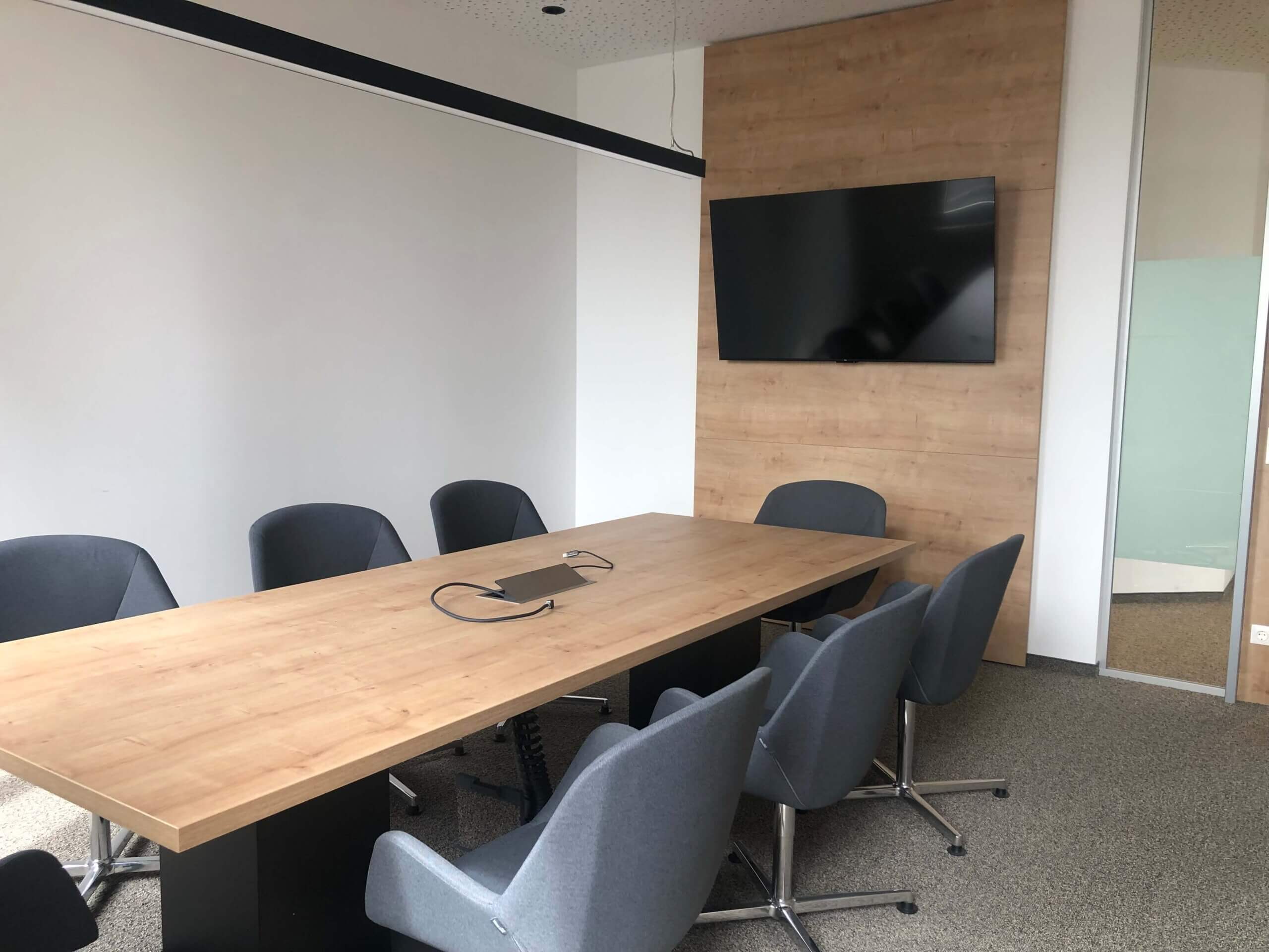 Ein kleiner Konferenzraum in der Raiffeisenbank Oberwart. Ein rechteckiger Holztisch ist von grauen Stühlen umgeben. An der Wand hinter dem Tisch hängt ein großer Flachbildfernseher. Die Wände sind weiß, und der Raum hat eine schlichte, moderne Einrichtung.