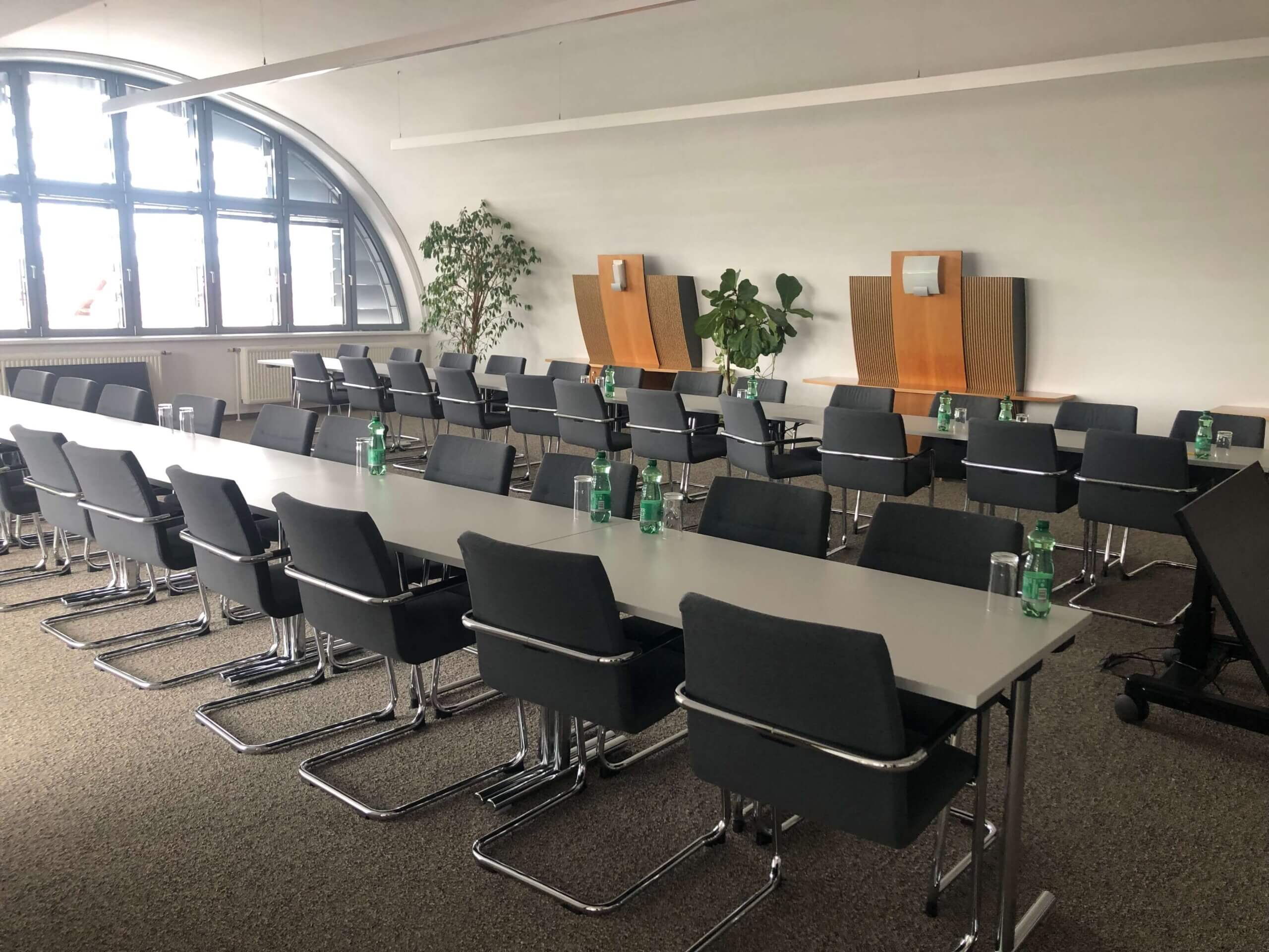 Ein heller Konferenzraum in der Raiffeisenbank Oberwart. Lange Tische sind in zwei Reihen aufgestellt, bestückt mit dunkelgrauen Stühlen. Auf den Tischen stehen grüne Wasserflaschen und Gläser. Links ist ein großes Bogenfenster, das den Raum mit natürlichem Licht durchflutet. An der Wand sind dekorative Holzelemente und Pflanzen.