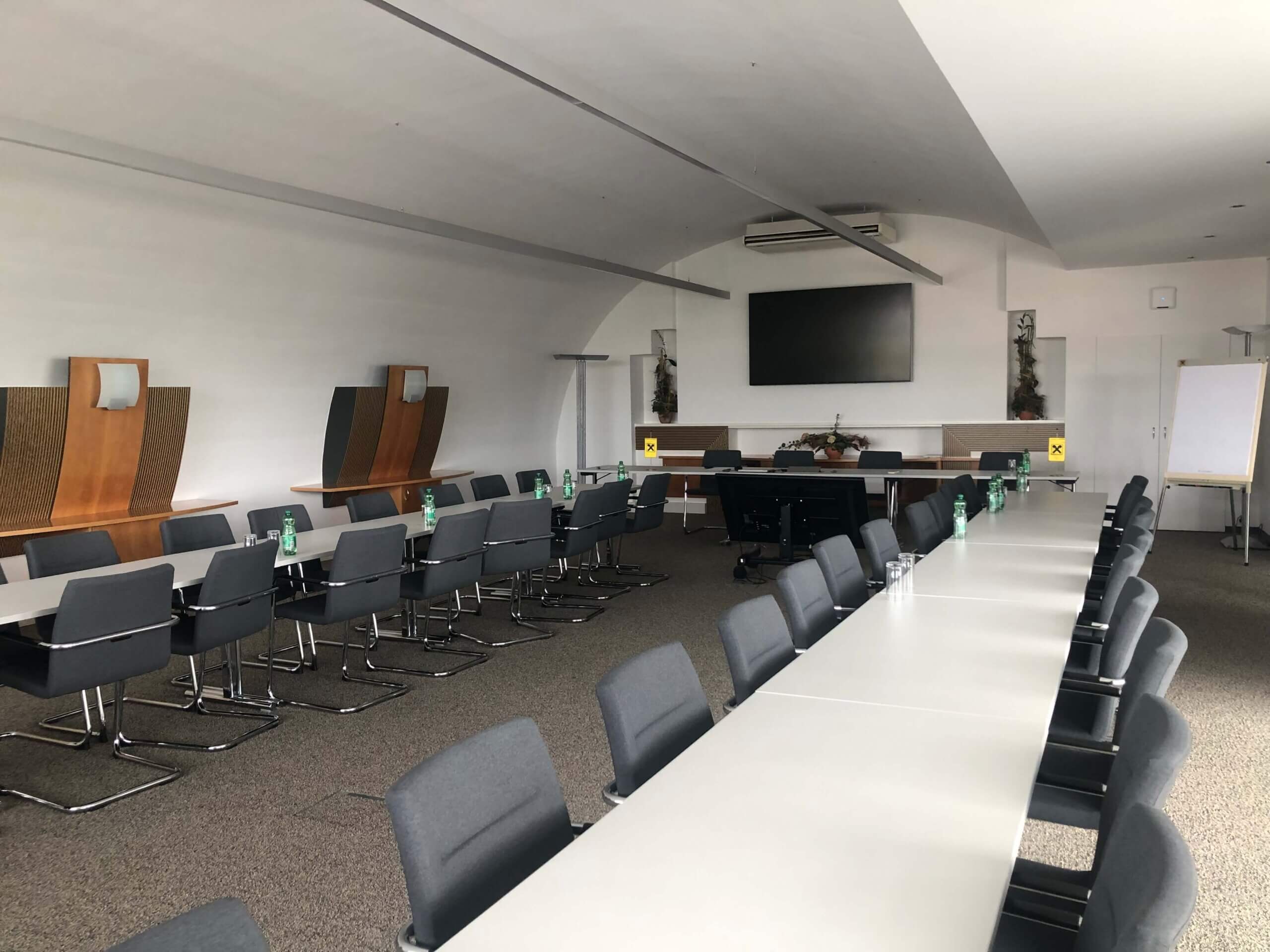 Ein weiterer Blick auf den Konferenzraum in der Raiffeisenbank Oberwart. Die Tische und Stühle sind aus einer anderen Perspektive zu sehen. Am Ende des Raums hängt ein großer Flachbildfernseher an der Wand. Auf den Tischen stehen weiterhin grüne Wasserflaschen und Gläser. Links steht ein Whiteboard, und im Raum befinden sich mehrere Pflanzen.