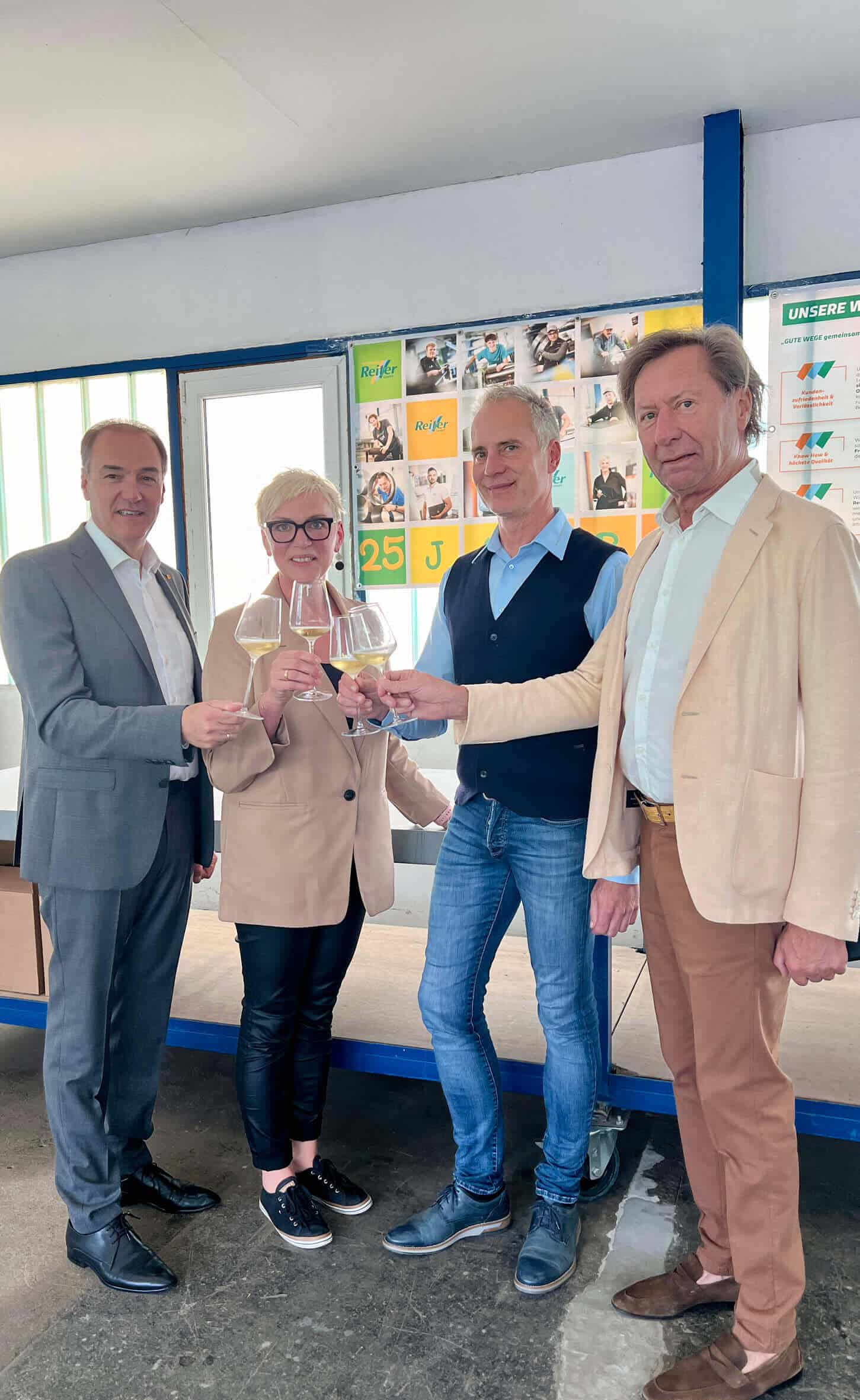 Das Bild zeigt vier Personen in einem hell beleuchteten Raum, die gemeinsam mit Weingläsern anstoßen. Landesrat Leonhard Schneemann steht links, gekleidet in einem grauen Anzug und einem weißen Hemd ohne Krawatte, und lächelt, während er ein Weinglas hält. Neben ihm steht Johanna Reiter und dann David Reiter, der eine blaue Weste über einem hellblauen Hemd und dunkelblaue Jeans trägt. Er steht zwischen seiner Frau und dem Landesrat und hält ebenfalls ein Weinglas. Johanna Reiter trägt eine beige Jacke über einem schwarzen Top und schwarze Lederhosen. Sie steht neben ihrem Mann und lächelt, während sie mit den anderen anstößt. Ganz rechts im Bild steht Kurt Maczek, der Bürgermeister von Pinkafeld, gekleidet in ein hellbeiges Sakko und eine braune Hose, auch er hält ein Weinglas und sieht in die Kamera. Im Hintergrund ist eine Wand mit Bildern und Plakaten zu sehen, die verschiedene Aktivitäten und Meilensteine der Firma Reiter im Laufe der Jahre zeigen, darunter ein Plakat, das „25 J“ vermerkt, um das 25-jährige Firmenjubiläum zu kennzeichnen. Die Stimmung im Bild wirkt feierlich und freundlich.