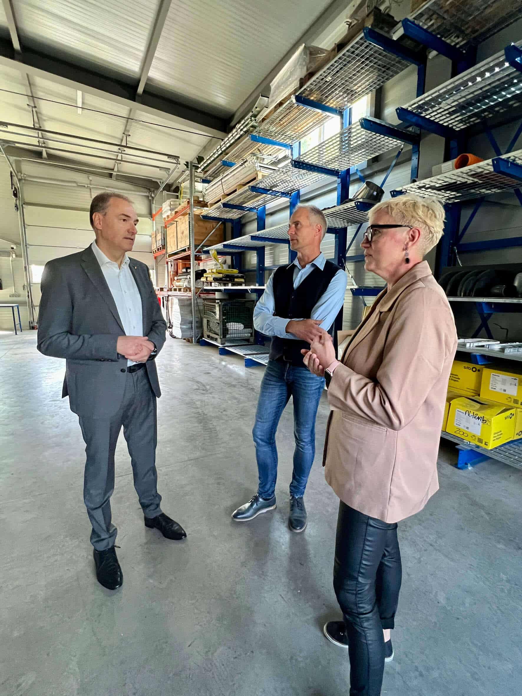 Das Bild zeigt drei Personen in einer Lagerhalle, die in einer Diskussion vertieft sind. Links steht Landesrat Leonhard Schneemann, gekleidet in einem dunkelgrauen Anzug und einem weißen Hemd ohne Krawatte. Er hört aufmerksam zu, sein Kopf leicht geneigt. In der Mitte befindet sich der Unternehmer David Reiter, leger gekleidet in einer blauen Weste über einem hellblauen Hemd und dunklen Jeans, die Arme verschränkt, während er das Gespräch führt. Rechts steht seine Frau Johanna Reiter, die eine beige Jacke über einem schwarzen Top und schwarzen Lederhosen trägt. Sie hat kurze blonde Haare, trägt eine Brille und hat ihre Hände ineinander gelegt, während sie aufmerksam den anderen beiden zuhört. Im Hintergrund sind mehrere Regale mit gestapelten Materialien und Werkzeugen sichtbar. Die Umgebung wirkt sauber und gut organisiert, typisch für eine industrielle Lagerhalle. Die Beleuchtung im Raum ist hell und gleichmäßig.