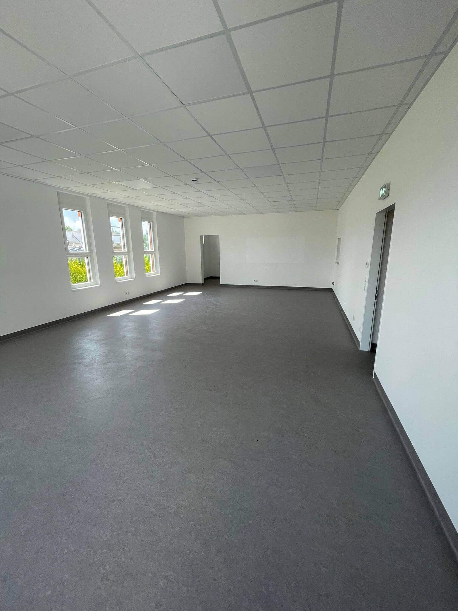 Ein großer, heller Raum mit mehreren Fenstern, die viel Licht hereinlassen. Die Wände sind weiß gestrichen und der Boden ist grau. Die Decke hat kleine Löcher und moderne Lichtleisten. Der Raum ist leer und bietet viel Platz für zukünftige Aktivitäten.