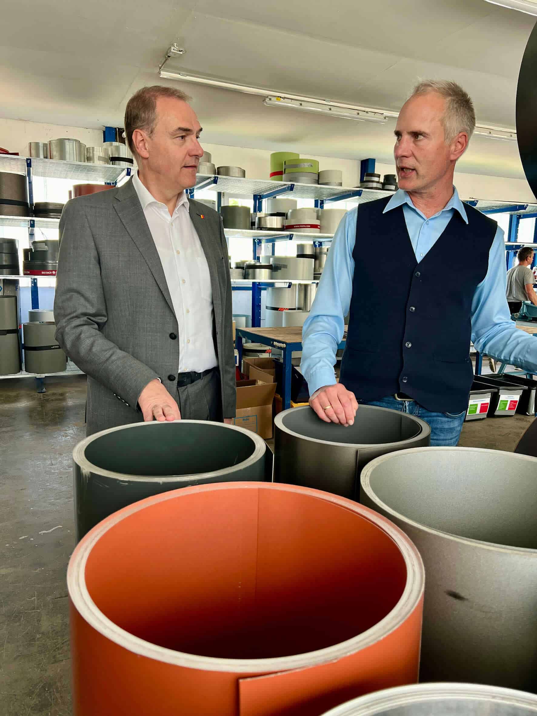 Das Bild zeigt zwei Männer, die in einer industriellen Umgebung, vermutlich in einem Lager oder einer Produktionsstätte, stehen und miteinander sprechen. Auf der linken Seite steht Landesrat Leonhard Schneemann. Er ist gekleidet in einen grauen Anzug, ein weißes Hemd ohne Krawatte und trägt ein Abzeichen am Revers. Sein Ausdruck ist aufmerksam und er blickt in Richtung des anderen Mannes. Auf der rechten Seite steht der Unternehmer Dr. Mag. David Reiter. Er trägt eine blaue Weste über einem hellblauen Hemd und eine dunkle Hose. Sein Gesichtsausdruck wirkt engagiert, während er spricht und gestikuliert. Im Hintergrund des Bildes sind mehrere große, zylindrische Objekte in verschiedenen Farben zu sehen, die auf Regalen gelagert werden. Die Szene ist in einem Raum mit hellen Wänden und Deckenleuchten aufgenommen, was eine klare und gut beleuchtete Umgebung schafft. Beide Männer wirken in ein ernsthaftes Gespräch vertieft, das sich um die sichtbaren industriellen Materialien oder Produkte zu drehen scheint.