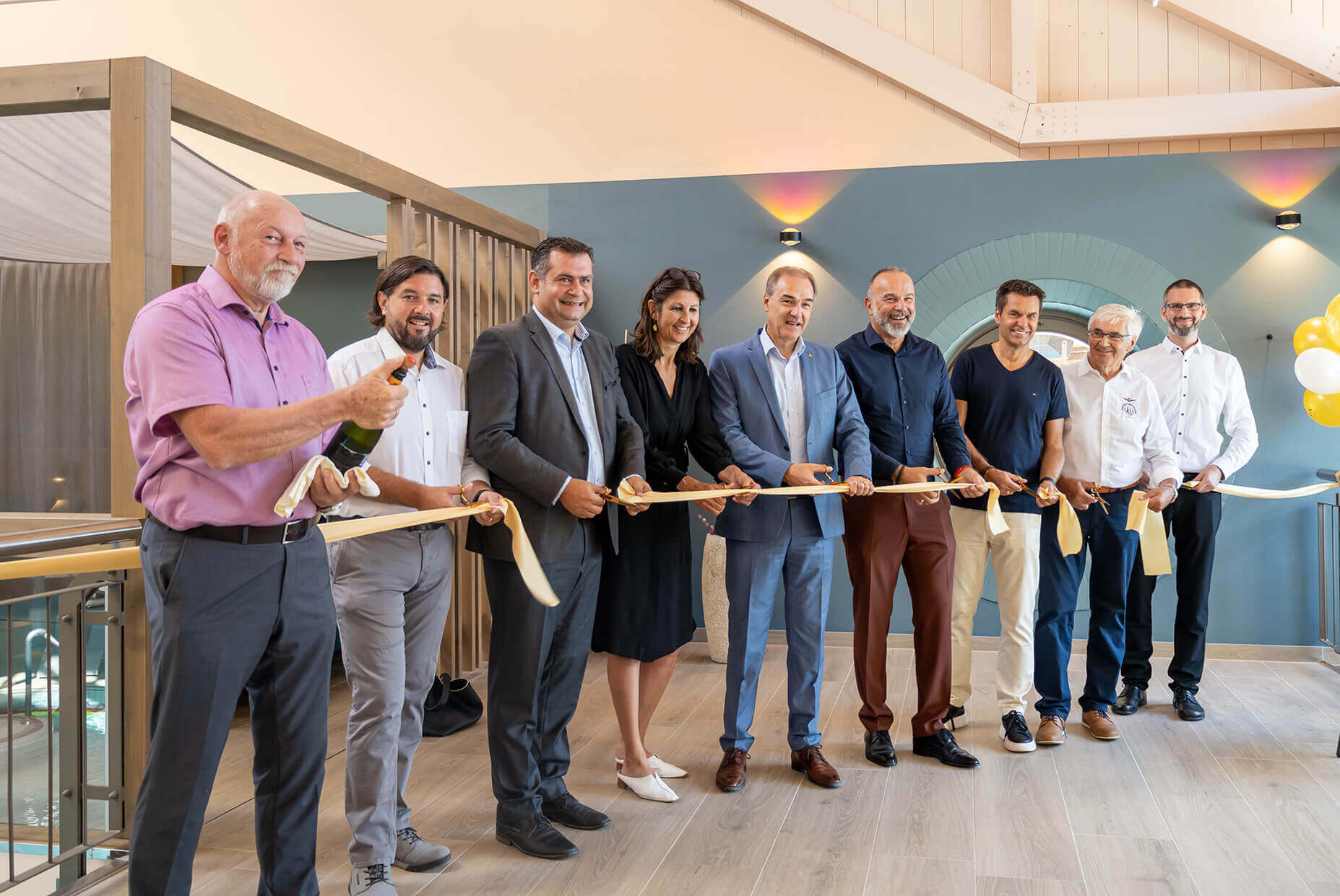 Feierliche Eröffnung der Avita Therme: Bürgermeister Hans Unger (links)steht neben einem Herren, der eine geöffnete Champagnerflasche hält und lächelt, während neben ihm Bürgermeister Stefan Laimer steht. Architektin Sylvia Benno-Zembacher, Landesrat Leonhard Schneemann, Avita-Geschäftsführer Peter Prisching, Burgenland Tourismus Geschäftsführer Didi Tunkel und Kommanditistenvertreter Ewald Töilly stehen neben ihm in einer Reihe. Alle halten gemeinsam ein goldenes Band, das zur Eröffnung durchgeschnitten wird. Im Hintergrund sind helle Holzkonstruktionen und eine blau-graue Wand zu sehen. Auf der rechten Seite sind gelbe und weiße Luftballons angebracht. Foto© Avita Resort