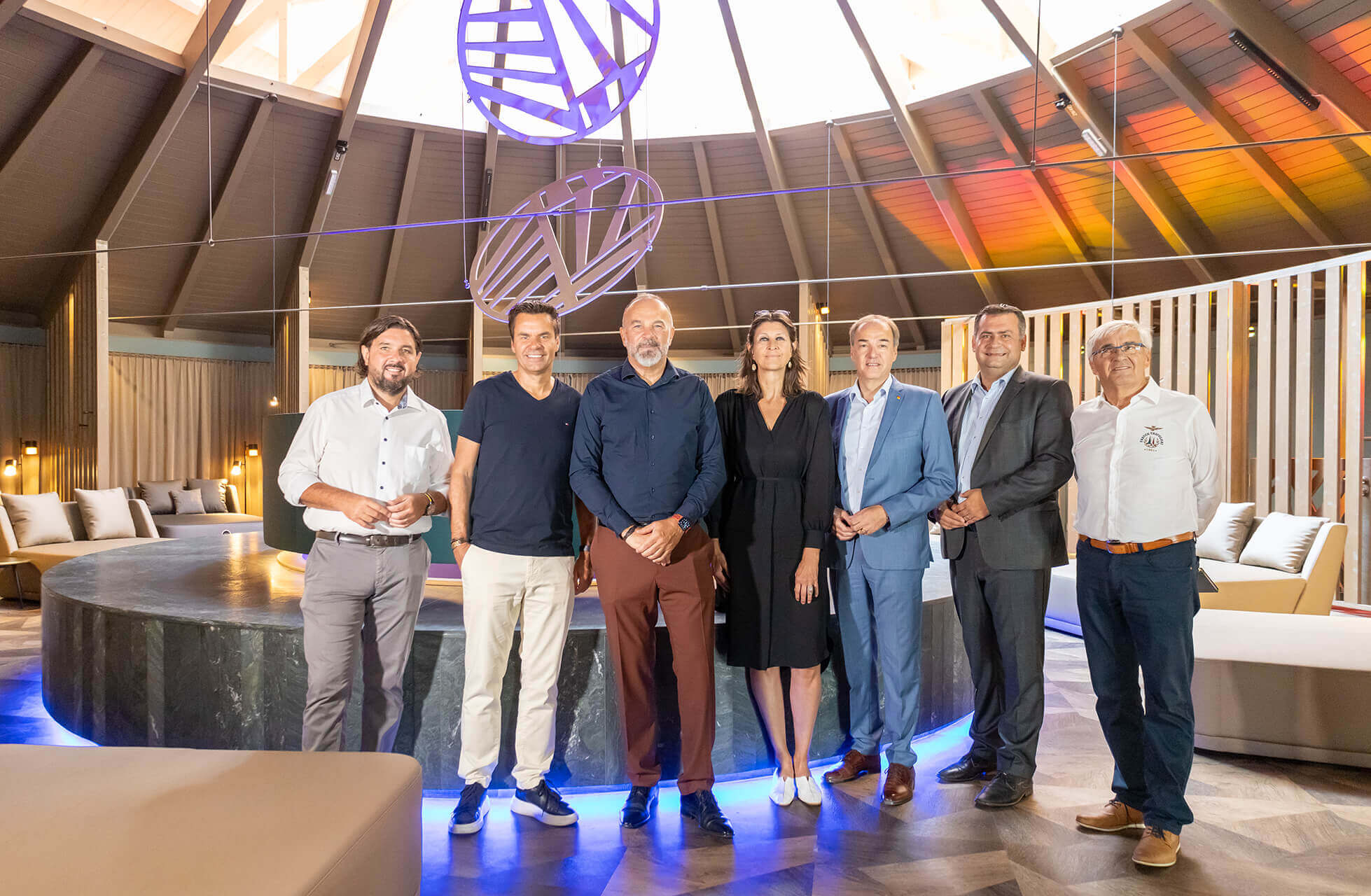 Bürgermeister Stefan Laimer, Geschäftsführer Didi Tunkel, Geschäftsführer Peter Prisching, Landesrat Leonhard Schneemann, Architektin Sylvia Benno-Zembacher, Bürgermeister Hans Unger und Kommanditistenvertreter Ewald Tölly (von links nach rechts) stehen in einem eleganten Raum mit hoher, gewölbter Decke. Der Raum ist modern gestaltet mit beigen Sitzgelegenheiten und einem großen, runden Tisch in der Mitte, der von blauen Lichtern beleuchtet wird. Über ihnen hängen zwei große, leuchtende Kunstinstallationen. Die Personen lächeln und stehen entspannt nebeneinander. Im Hintergrund sind Vorhänge und moderne Lampen zu sehen.©Avita Resort