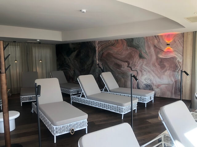 Ein Ruheraum im Spa ist mit mehreren hellgrauen Liegestühlen ausgestattet, die in Reihen auf einem dunklen Holzboden angeordnet sind. Die Wände sind mit einem großen, abstrakten Wandgemälde in Wirbeln aus Rosa, Grün und Schwarz verziert. Sanfte Beleuchtung sorgt für ein warmes Licht in der ruhigen Umgebung. Vorhänge bedecken teilweise die Rückwand und tragen zur entspannten Atmosphäre bei.