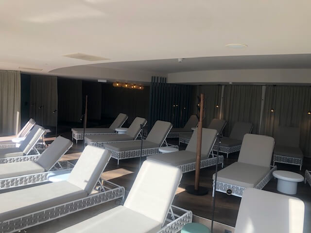 Ein Ruheraum im Spa ist mit mehreren hellgrauen Liegestühlen ausgestattet, die in Reihen auf einem dunklen Holzboden angeordnet sind. Die Wände sind mit einem großen, abstrakten Wandgemälde in Wirbeln aus Rosa, Grün und Schwarz verziert. Sanfte Beleuchtung sorgt für ein warmes Licht in der ruhigen Umgebung. Vorhänge bedecken teilweise die Rückwand und tragen zur entspannten Atmosphäre bei.