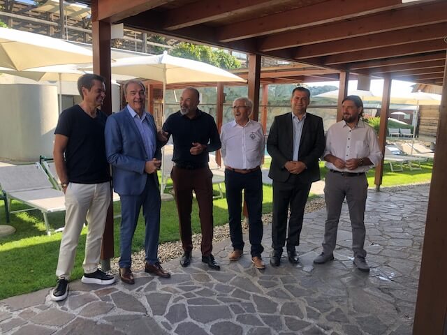Sechs Männer stehen in einem überdachten Bereich mit einem Boden aus Natursteinfliesen. Von links nach rechts tragen sie unterschiedlich formelle Kleidung: Der erste Mann trägt ein schwarzes T-Shirt und helle Hosen, der zweite einen blauen Anzug ohne Krawatte, der dritte ein schwarzes Hemd und dunkle Hosen, der vierte ein weißes Hemd mit einem Logo und dunkle Hosen, der fünfte einen dunklen Anzug, und der sechste ein weißes Hemd mit hellen Hosen. Im Hintergrund sind Liegestühle und Sonnenschirme auf einer Wiese sowie eine Pergola und weitere Gebäudeteile zu sehen.