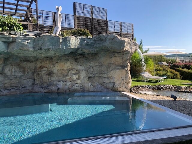 Ein Außenpool mit klarem, blauem Wasser liegt vor einer Felsformation, die eine natürliche Klippe nachbildet. Auf der Klippe steht eine moderne, weiße Skulptur in menschlicher Form. Im Hintergrund sind eine Pergola und grüne Pflanzen zu sehen. Rechts vom Pool befinden sich Liegestühle unter weißen Sonnenschirmen auf einer gepflegten Wiese. Der Himmel ist klar und blau, was zu der friedlichen Atmosphäre beiträgt.