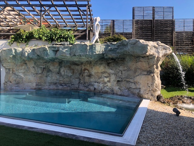 Ein Außenpool mit klarem, blauem Wasser liegt vor einer Felsformation, die eine natürliche Klippe nachbildet. Auf der Klippe steht eine moderne, weiße Skulptur in menschlicher Form. Im Hintergrund sind eine Pergola und grüne Pflanzen zu sehen. Rechts vom Pool befinden sich Liegestühle unter weißen Sonnenschirmen auf einer gepflegten Wiese. Der Himmel ist klar und blau, was zu der friedlichen Atmosphäre beiträgt.