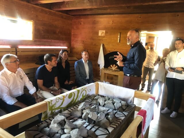 Eine Gruppe von sieben Personen befindet sich in einer Sauna mit Holzwänden und -bänken. Der Mann rechts im schwarzen Hemd steht und spricht zu den anderen, die sitzen und aufmerksam zuhören. Zwei Frauen und vier Männer sind zu sehen, alle in unterschiedlicher Kleidung, von Freizeitkleidung bis hin zu formellen Anzügen. In der Mitte des Raumes befindet sich ein Saunaofen mit Steinen, der von Handtüchern und einem grünen Tuch umgeben ist. Tageslicht fällt durch ein kleines Fenster und erhellt den Raum.