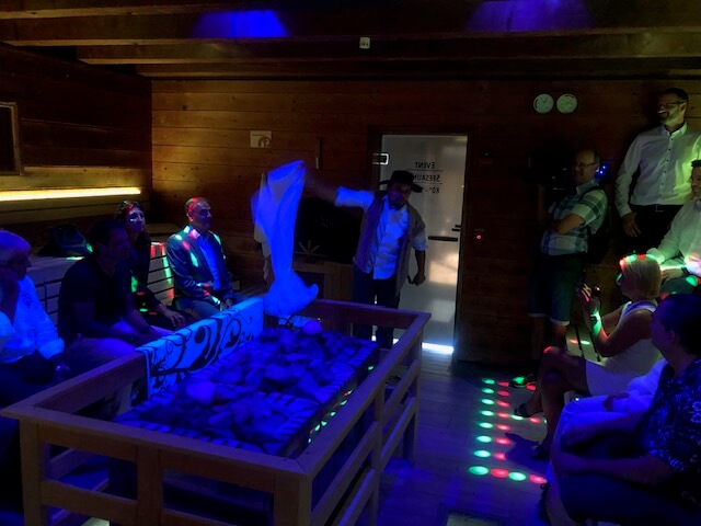 Eine Gruppe von neun Personen befindet sich in einer Sauna mit Holzwänden und -bänken. Die Sauna ist mit bunten, punktförmigen Lichtern beleuchtet, die eine festliche Atmosphäre schaffen. Ein Mann steht und wedelt ein weißes Handtuch, während die anderen auf den Bänken sitzen und zuschauen. In der Mitte des Raumes befindet sich ein Saunaofen mit Steinen, der von Handtüchern und einem dekorativen Tuch umgeben ist. Zwei Kameraleute filmen die Szene. Durch die geöffnete Tür fällt Tageslicht herein.