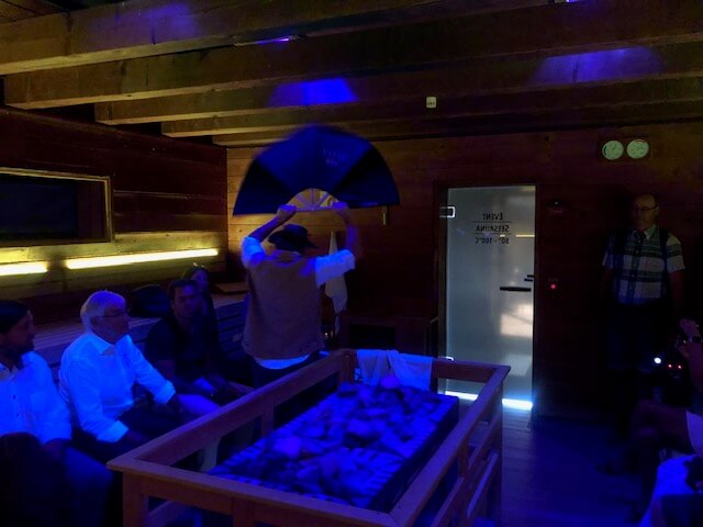 Eine Gruppe von neun Personen befindet sich in einer Sauna mit Holzwänden und -bänken. Die Sauna ist mit bunten, punktförmigen Lichtern beleuchtet, die eine festliche Atmosphäre schaffen. Ein Mann steht und wedelt ein weißes Handtuch, während die anderen auf den Bänken sitzen und zuschauen. In der Mitte des Raumes befindet sich ein Saunaofen mit Steinen, der von Handtüchern und einem dekorativen Tuch umgeben ist. Zwei Kameraleute filmen die Szene. Durch die geöffnete Tür fällt Tageslicht herein.