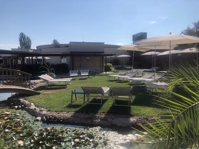 Ein Außenbereich eines Spas zeigt mehrere hellgraue Liegestühle, die auf einer gepflegten Rasenfläche unter weißen Sonnenschirmen stehen. Links ist eine kleine Holzbrücke über einen Teich zu sehen, der mit Seerosen bedeckt ist. Im Hintergrund befindet sich ein modernes Gebäude mit flachem Dach, umgeben von weiteren Grünflächen und Bäumen. Das Wetter ist sonnig, und der Himmel ist klar und blau.