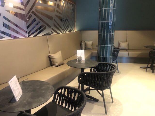 Ein gemütlicher Lounge-Bereich mit beigen Polsterbänken, die entlang der Wände verlaufen. Kleine runde Tische mit schwarzen Stühlen stehen vor den Bänken. An den Tischen liegen Menükarten. Eine große Säule, verkleidet mit grünen, glänzenden Fliesen, steht im Raum. Die Wände sind in einem dunklen Grünton gestrichen, und ein Fenster mit modernem, geometrischem Muster aus Milchglas trennt diesen Bereich von einem anderen Raum. Dezente Beleuchtung schafft eine entspannte Atmosphäre.