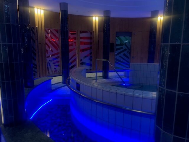 Ein Wellnessbereich mit mehreren runden, gefliesten Becken, die durch eine blaue Unterwasserbeleuchtung stimmungsvoll in Szene gesetzt sind. Die Wände sind aus Holz mit großen, geometrisch gemusterten Milchglasscheiben in verschiedenen Farben. Dunkel geflieste Säulen verlaufen bis zur Decke und strukturieren den Raum. Eine Stange und Stufen führen in eines der Becken. Die Beleuchtung ist gedämpft und trägt zu einer beruhigenden Atmosphäre bei. Steine sind im Bodenbereich rund um die Becken zu sehen, was das natürliche Ambiente verstärkt.