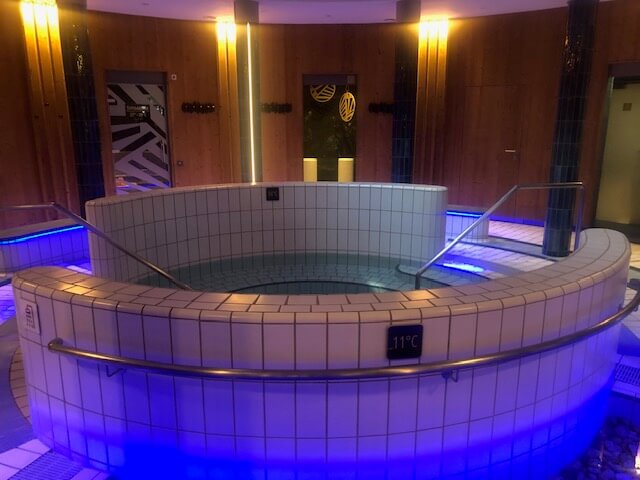 Ein runder Whirlpool mit weißen Fliesen und blauer Unterwasserbeleuchtung dominiert diesen Wellnessbereich. Zwei Handläufe führen in das Becken, das mit einem Schild mit der Temperaturanzeige "11°C" versehen ist. Die Wände sind mit Holz verkleidet und mit geometrisch gemusterten Milchglasscheiben in verschiedenen Farben dekoriert. Mehrere dunkel geflieste Säulen tragen zur Struktur des Raumes bei. Gedämpfte, warme Beleuchtung sorgt für eine beruhigende Atmosphäre, die durch die klaren Linien und das moderne Design betont wird.