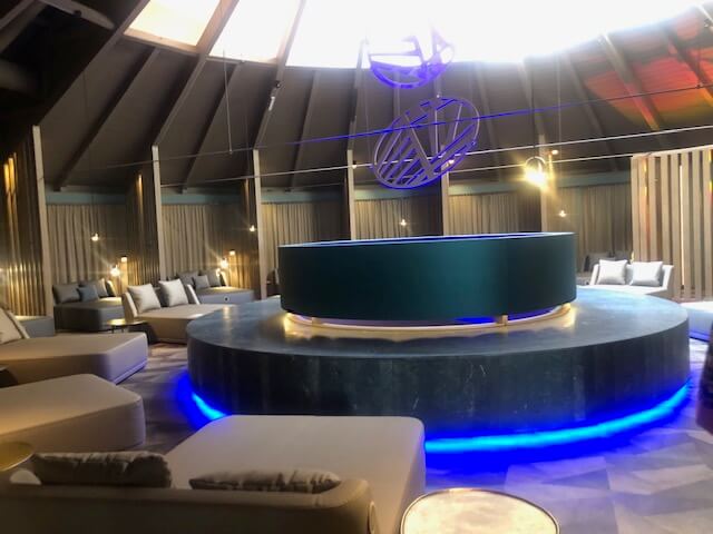 Ein eleganter Lounge-Bereich in einem Spa mit einem großen, runden Sitzbereich in der Mitte, der von blauer Unterbeleuchtung akzentuiert wird. Der Raum hat eine hohe, kegelförmige Decke mit einer großen Öffnung, durch die Tageslicht einfällt. Um die zentrale Sitzgruppe herum sind bequeme Liegen mit hellen Polstern und Kissen verteilt. Die Wände sind mit vertikalen Holzpaneelen verkleidet, und dezente Beleuchtung schafft eine entspannte Atmosphäre. An der Decke hängen moderne, geometrische Skulpturen, die dem Raum ein stilvolles und zeitgemäßes Ambiente verleihen.