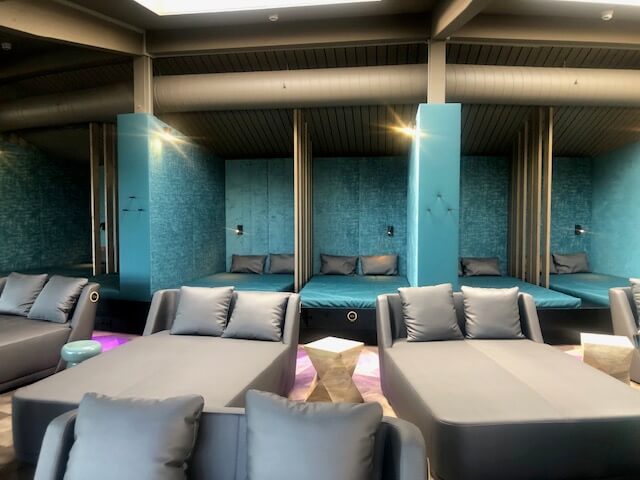 Ein moderner Entspannungsbereich in einem Spa mit mehreren Liegen und Sitzgelegenheiten in Grau- und Blautönen. Im hinteren Teil des Raumes befinden sich Nischen mit breiten, gepolsterten Liegen, die durch türkisfarbene Wände und vertikale Holzpaneele voneinander getrennt sind. Vor diesen Nischen stehen graue Sofas und Liegen mit passenden Kissen. Der Boden ist mit einem modernen Muster gestaltet, und die Beleuchtung ist dezent und indirekt. Ein großes Belüftungsrohr verläuft entlang der Decke, was dem Raum ein industrielles Flair verleiht.