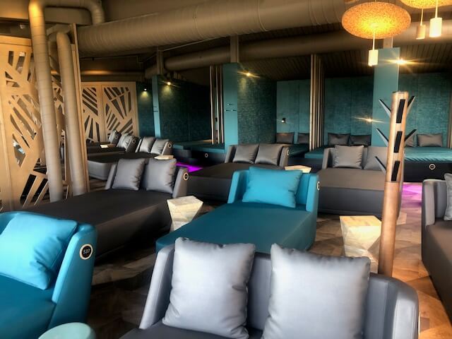 Ein moderner Entspannungsbereich in einem Spa mit mehreren Liegen und Sitzgelegenheiten in Grau- und Blautönen. Im hinteren Teil des Raumes befinden sich Nischen mit breiten, gepolsterten Liegen, die durch türkisfarbene Wände und vertikale Holzpaneele voneinander getrennt sind. Vor diesen Nischen stehen graue Sofas und Liegen mit passenden Kissen. Der Boden ist mit einem modernen Muster gestaltet, und die Beleuchtung ist dezent und indirekt. Ein großes Belüftungsrohr verläuft entlang der Decke, was dem Raum ein industrielles Flair verleiht.