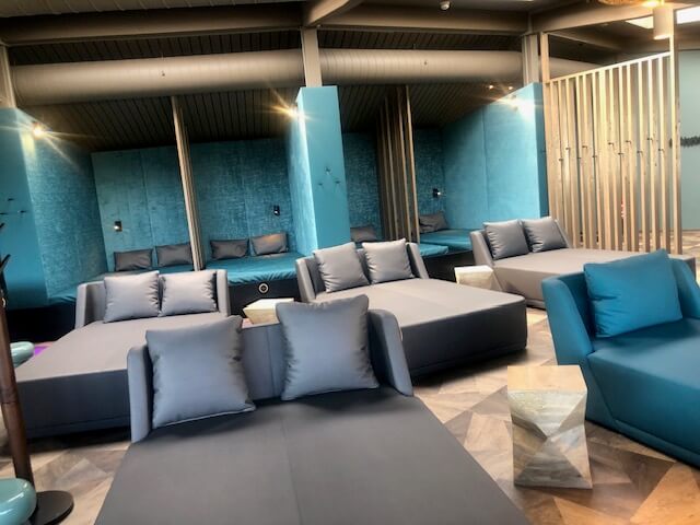 Ein moderner Entspannungsbereich in einem Spa mit mehreren Liegen und Sitzgelegenheiten in Grau- und Blautönen. Im hinteren Teil des Raumes befinden sich Nischen mit breiten, gepolsterten Liegen, die durch türkisfarbene Wände und vertikale Holzpaneele voneinander getrennt sind. Vor diesen Nischen stehen graue Sofas und Liegen mit passenden Kissen. Der Boden ist mit einem modernen Muster gestaltet, und die Beleuchtung ist dezent und indirekt. Ein großes Belüftungsrohr verläuft entlang der Decke, was dem Raum ein industrielles Flair verleiht.