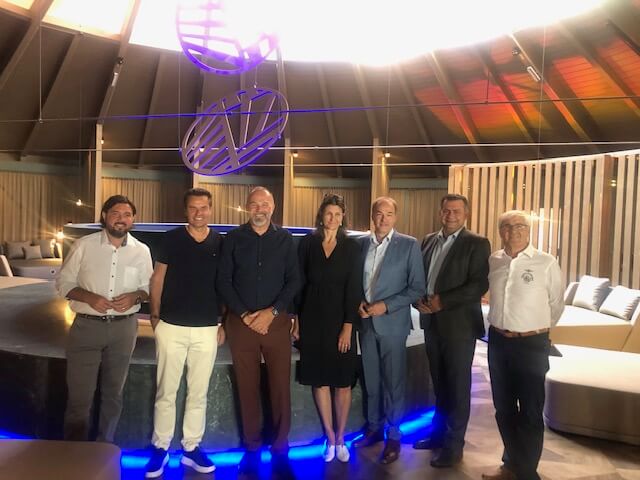 Bürgermeister Hans Unger, Burgenland Tourismusdirektor Didi Tunkel, Avita Geschäftsführer Peter Prisching, eine Architektin, Landesrat Leonhard Schneemann, Bürgermeister Stefan Lahmer und Kommanditistenvertreter Ewald Tölly stehen in einem modernen Lounge-Bereich. Sie sind unterschiedlich gekleidet: von Freizeitkleidung bis hin zu formellen Anzügen. Im Hintergrund sind runde Sitzbereiche mit blauer Unterbeleuchtung, eine hohe, kegelförmige Decke mit geometrischen Skulpturen und vertikale Holzpaneele an den Wänden zu sehen. Die Beleuchtung ist gedämpft und schafft eine stilvolle Atmosphäre.